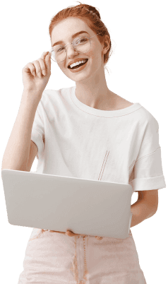 Image of a smiling, red haired woman standing with a laptop touching her glasses